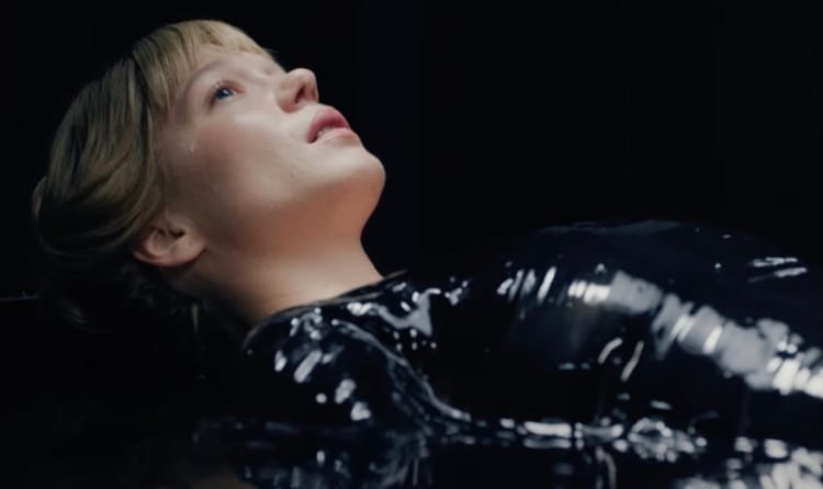 A blonde white woman in a black leather outfit lies in a pool of water. She is crying.