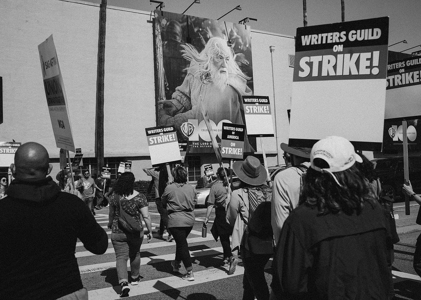 Striking writers and actors, carrying picket signs, cross the street outside Warner Bros. main studio, a giant image of Gandalf hovering over them.