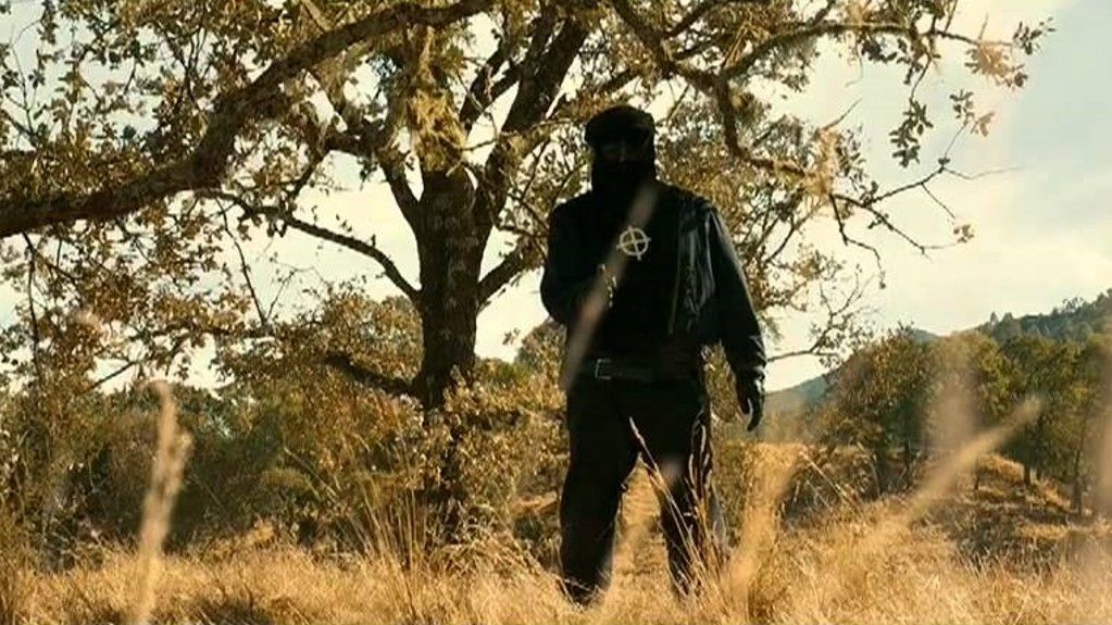 A man in all black, a Zodiac symbol on his chest, points a gun at someone off-camera.