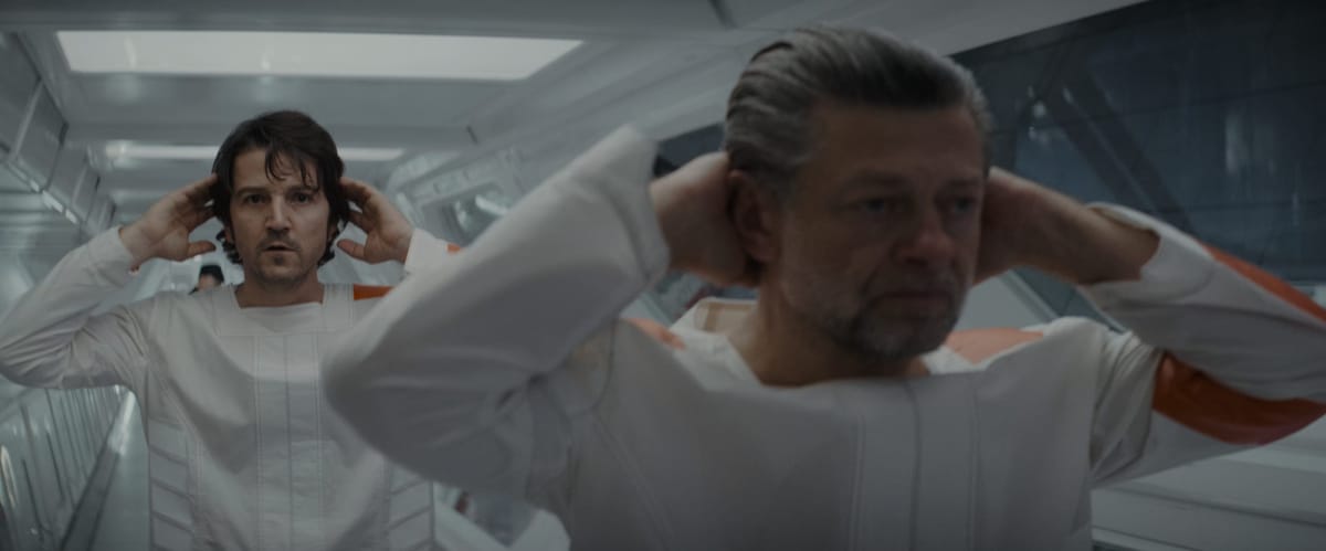 Cassian and Kino Loy stand at attention, hands behind their heads, in the hallway of the prison.