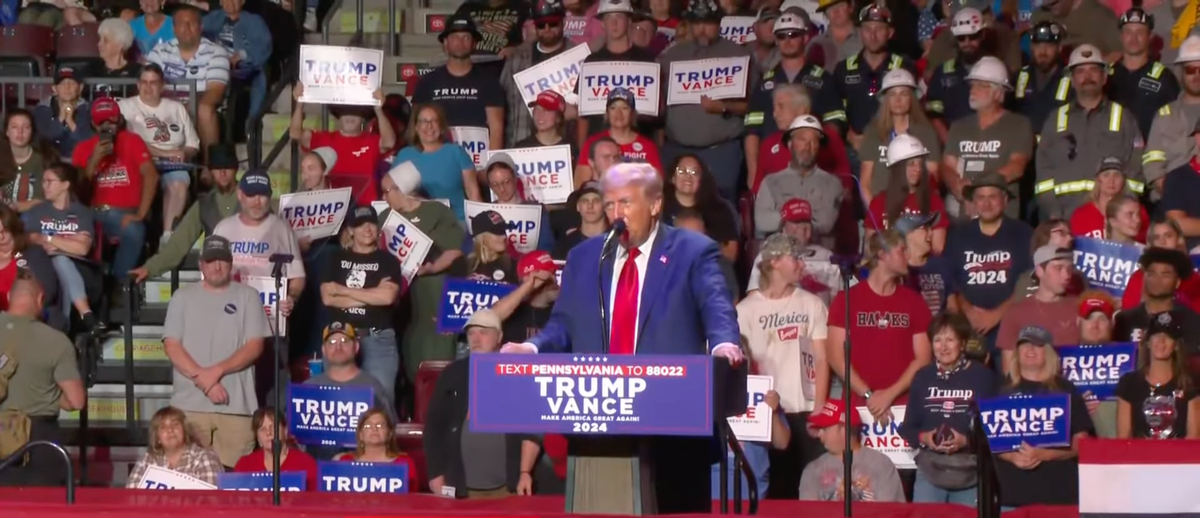 Donald Trump stands in front of a large crowd of supporters, speaking at a podium.