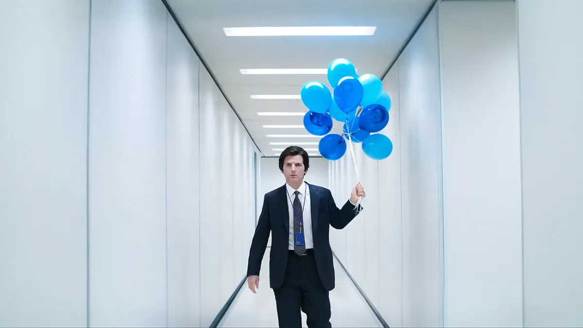 Mark S. (Adam Scott) is in a suit. He stands in a long, featureless, white hallway. He holds blue balloons with his face on them.