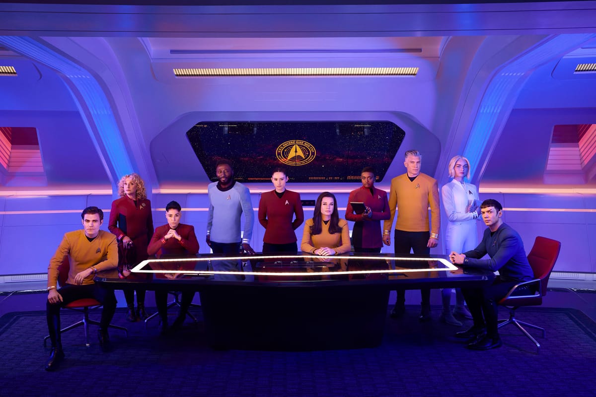 The cast of Star Trek: Strange New Worlds sits around a glowing table in a starship setting, wearing their Starfleet uniforms.