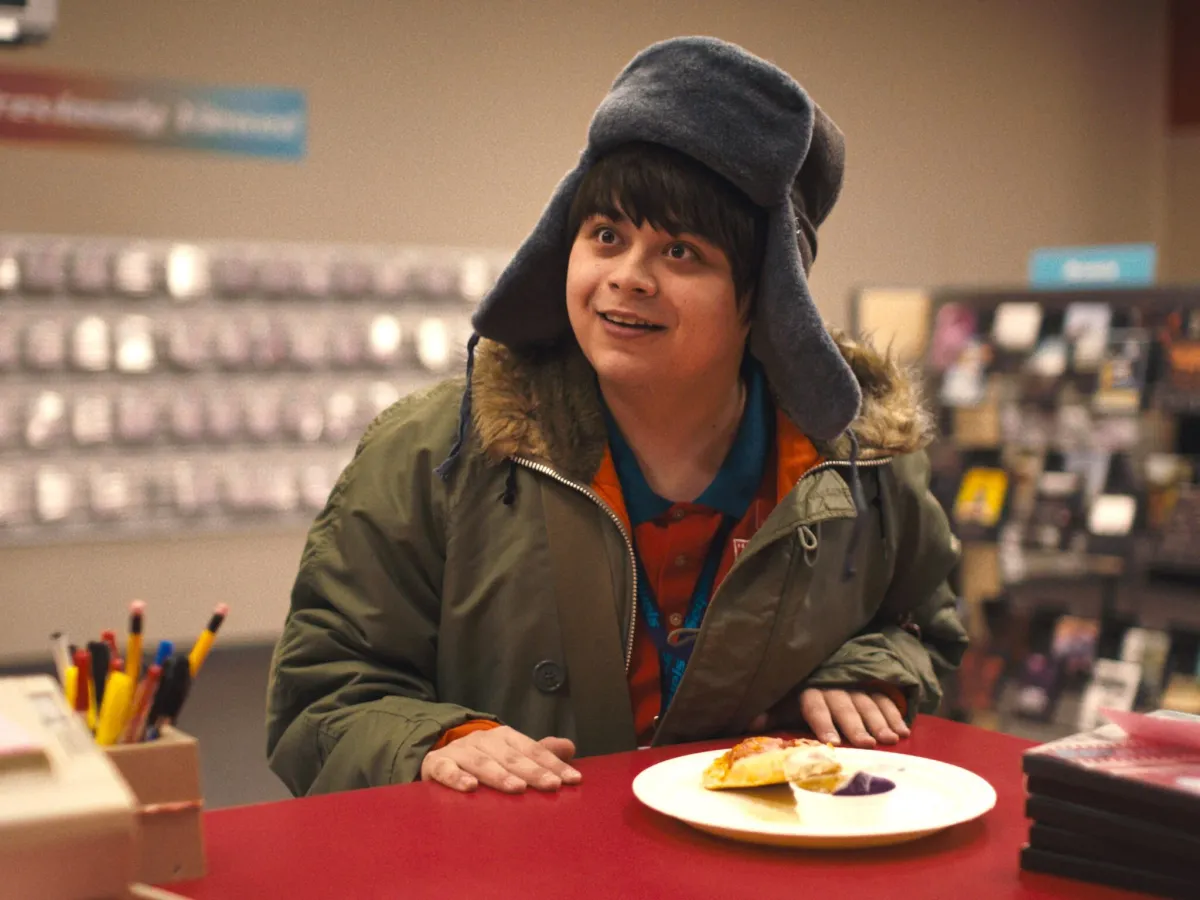 A white teenage boy wearing a ridiculous hat with earflaps is in a video store. A piece of pizza sits before him.