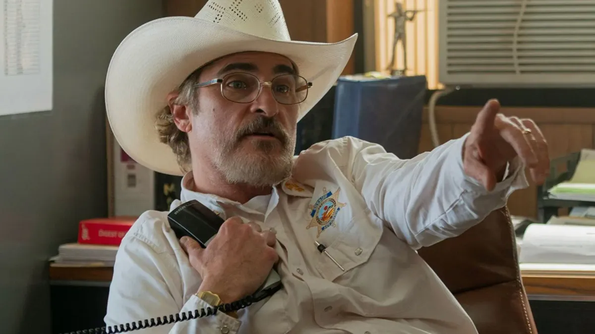 Joaquin Phoenix, in a white cowboy hat and white sheriff's shirt, holds a radio and points at someone off-camera.