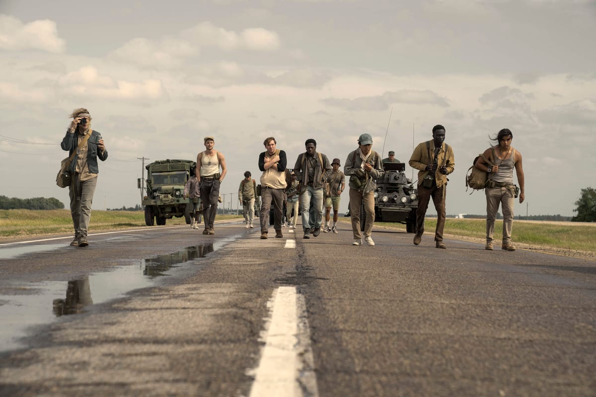 A group of young men walk down a long stretch of highway. They look absolutely exhausted. Men in military vehicles follow.