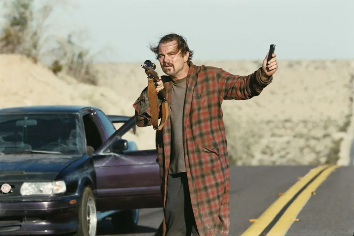 Leonardo DiCaprio, clad in a bathrobe and comfy clothes, stands on a desert highway, holding a gun, his beater car just behind him.
