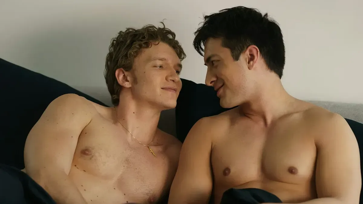 Ilya and Shane, both shirtless, lay together in bed and smile wistfully at one another.
