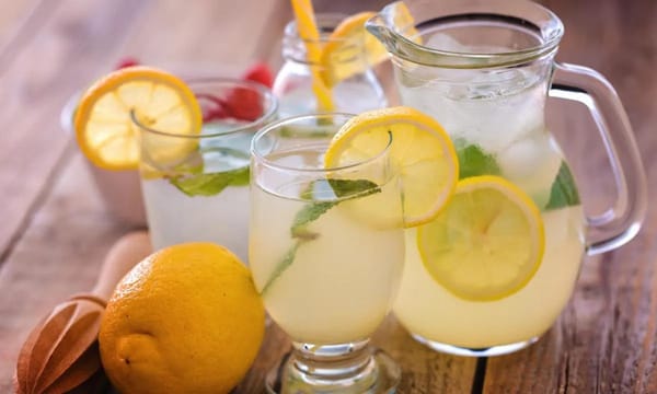 Ice cold refreshing lemonade, in several glasses and a large pitcher. Lemon slices and mint float in the drink.