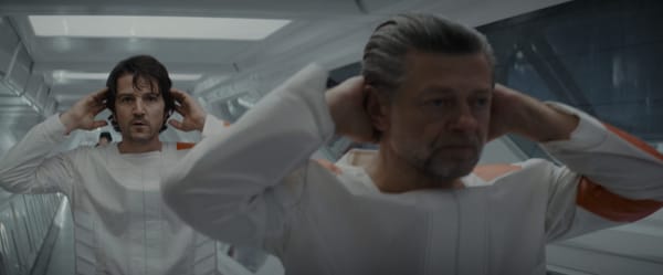 Cassian and Kino Loy stand at attention, hands behind their heads, in the hallway of the prison.