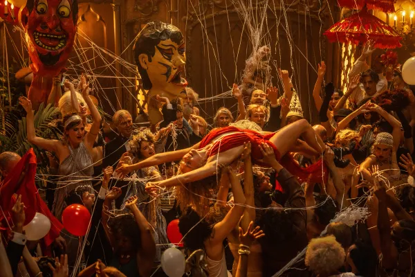 Margot Robbie, in a red dress, rides atop a throng of people, borne up by their hands, at a massive party.