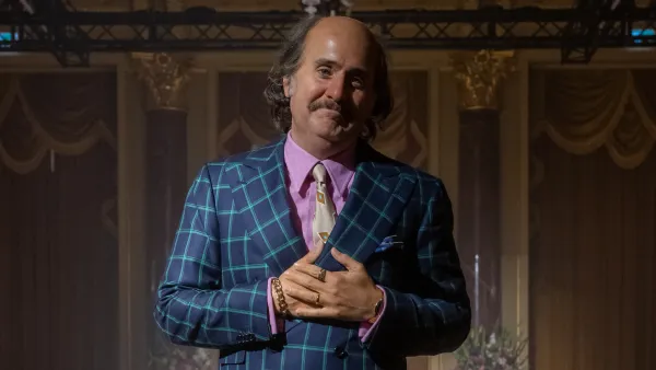 Jared Leto has a lot of padding added to his face via makeup in House of Guc. He wears a bald cap and a blue and green suit.