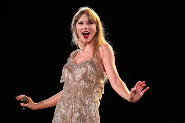 Taylor Swift, clad in a frilly gold dress, smiles as she performs in the Eras Tour concert. She's holding a microphone.