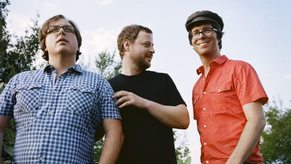 The members of Ben Folds Five pose in front of a forested area. Ben Folds looks at the camera; the other two look away.