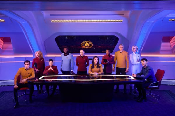 The cast of Star Trek: Strange New Worlds sits around a glowing table in a starship setting, wearing their Starfleet uniforms.