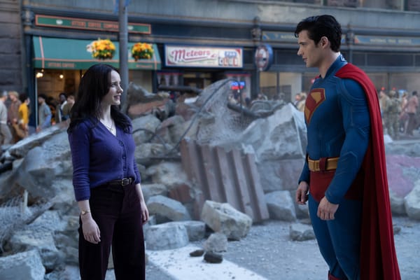 Lois Lane, brunette, white, blue eyes, wearing a purple sweater, regards Superman, white, brunette, in costume, amid post-battle rubble.