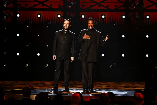 Lin-Manuel Miranda and Leslie Odom, Jr., both in all black suits, perform on a large stage.