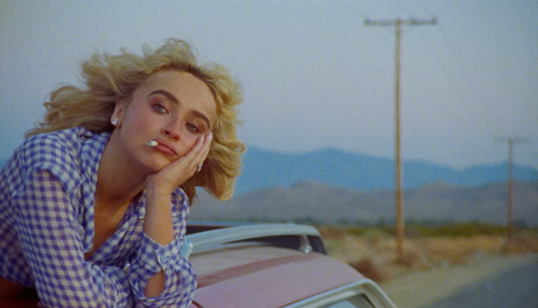 Dressed in a blue-white checkered top, Sabrina Carpenter hangs out of a car, cigarette in her mouth.