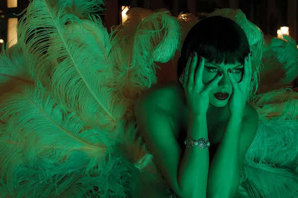 Taylor Swift, lit with a green light, wears an enormous feathered contraption and grips her face. She's got a dark brunette wig on.