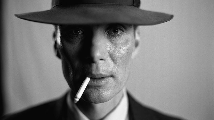 J. Robert Oppenheimer, played by Cillian Murphy, stares into the camera while smoking in this black-and-white image.