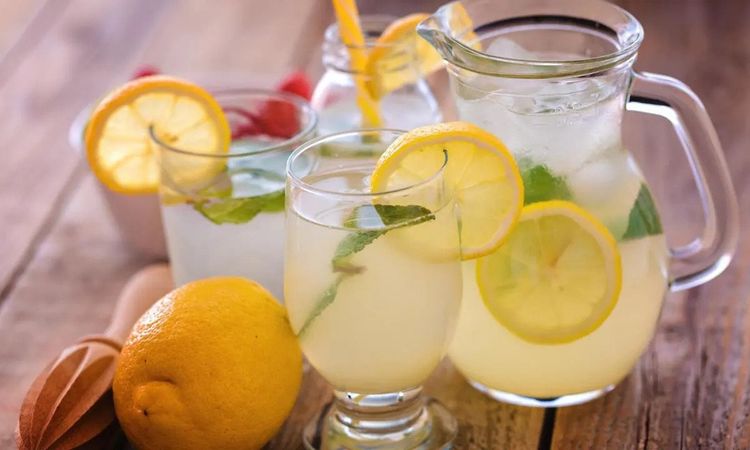 Ice cold refreshing lemonade, in several glasses and a large pitcher. Lemon slices and mint float in the drink.