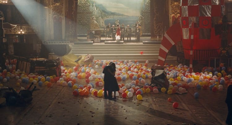 Two people embrace in the center of a ballroom, balloons having fallen on the floor all around them. A band plays onstage.