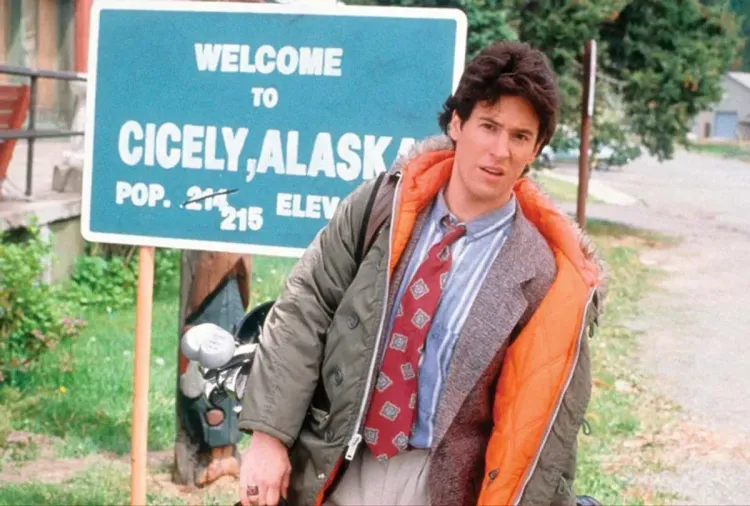 A man in a tie and parka looks befuddled while standing in front of a sign reading "Welcome to Cicely, Alaska. Pop. 215"