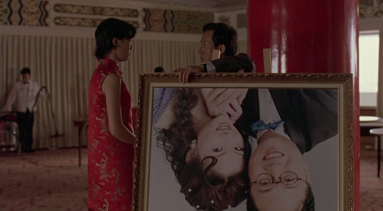 A girl talks to a man. They're both dressed in formalwear. The man leans on an upside-down photo of a couple.
