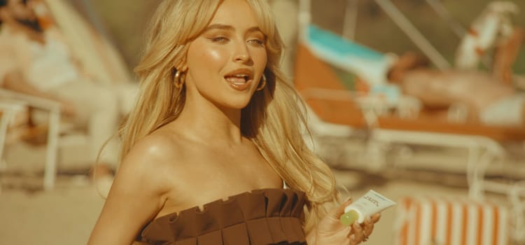 Sabrina Carpenter in the "Espresso" video, all blonde hair and golden skin, holding sunscreen.