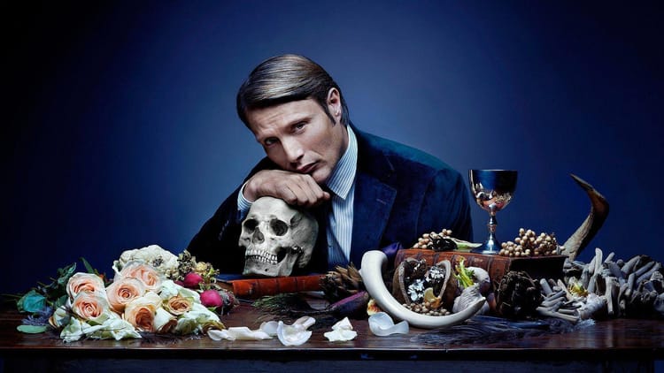 Hannibal (from the TV show) sits at a table covered in flowers, a skull, and other arcane symbols.