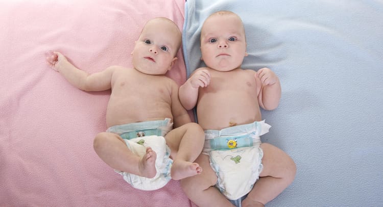 Two babies, both bald, both in diapers, lay on two blankets. One blanket is pink, the other blue.