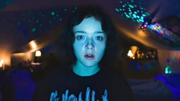 A pale girl with brown eyes stares directly into the camera. She is in a dark bedroom lit with small lamps and glow-in-thed