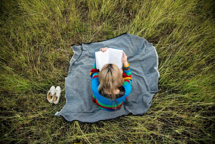 A view of a person with long blonde hair sitting on a blanket and reading. We see them from above.