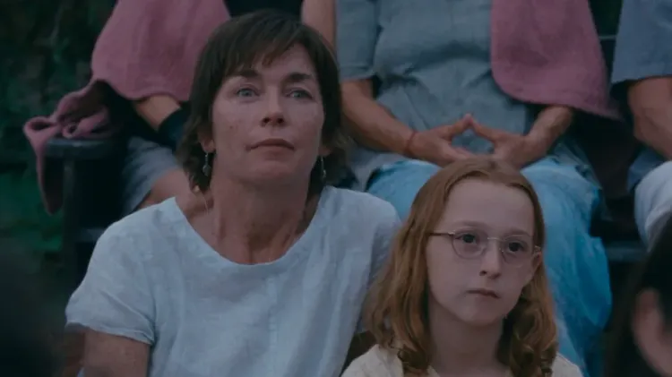 A short-haired woman in a white T-shirt sits with a red-haired girl in glasses. They are watching something.