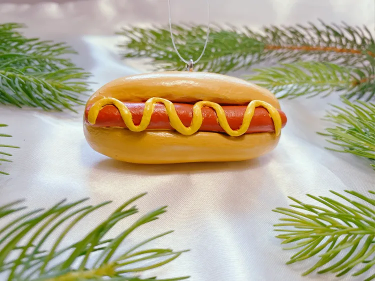 A small, plastic hot dog in a bun, covered in mustard, rests among pine branches. It has a little loop atop it for hangi