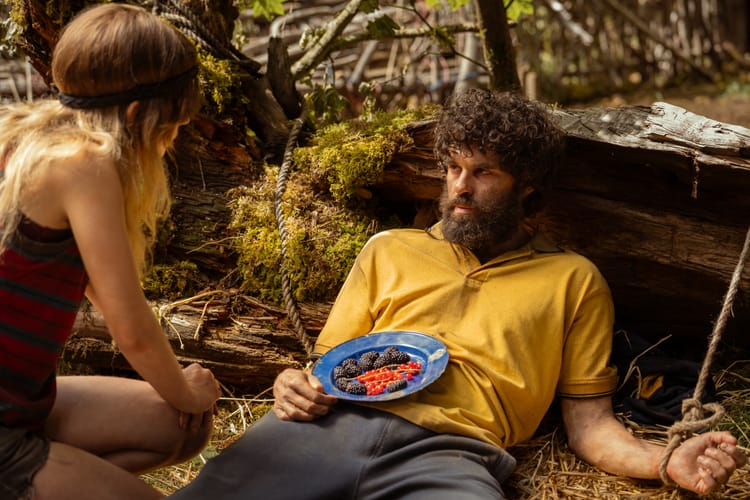 In a scene from Yellowjackets, Ben, who is tied down, looks up at Natalie. He has a plate of berries on his stomach.