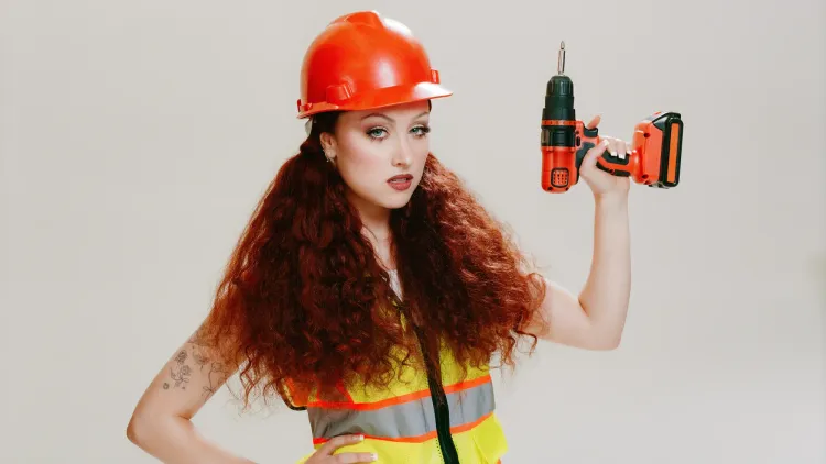 Chappell Roan in an orange hardhat and yellow reflective work vest, holding a power drill.