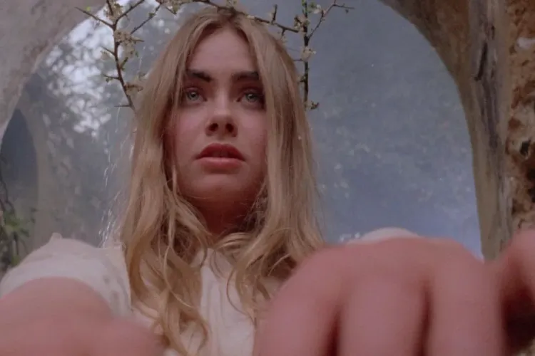 A white, blonde woman with blue eyes, clad all in white, wearing a crown of sticks, leans toward camera in close-up.