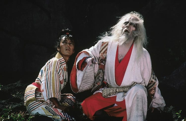 Two people crouch in the wilderness, one old, one young. They stare at the sky in horror. 
