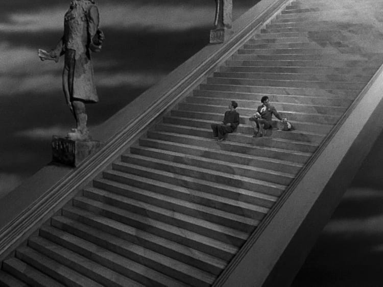Two people sit on an enormous, seemingly endless staircase, buttressed by giant statues, in an extreme wide shot.