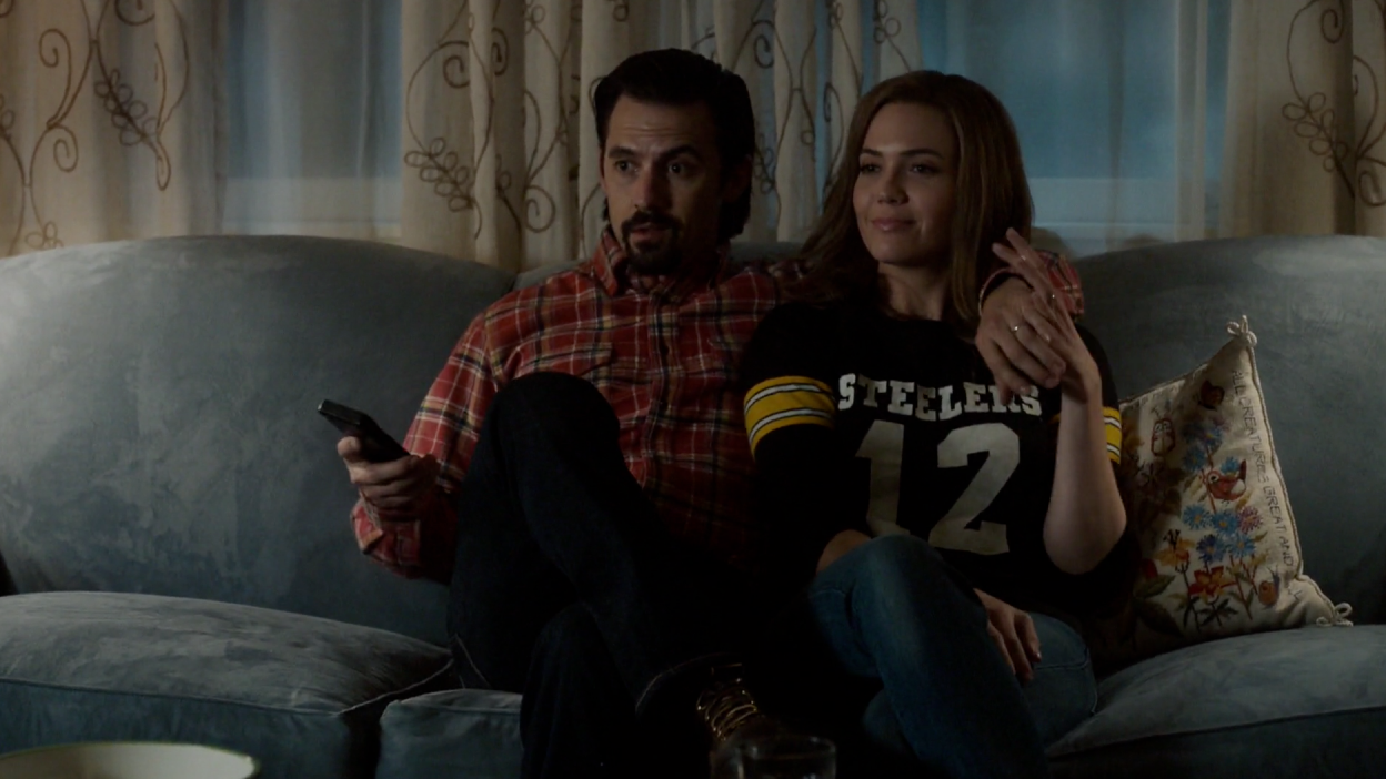 Milo Ventimiglia and Mandy Moore (in a Steelers jersey) watch some TV on the couch in This Is Us.