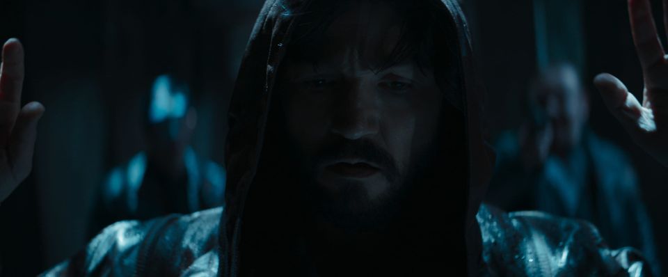 Cassian holds his hands aloft as two guards stop him. The guards are out of focus over Cassian's shoulders.