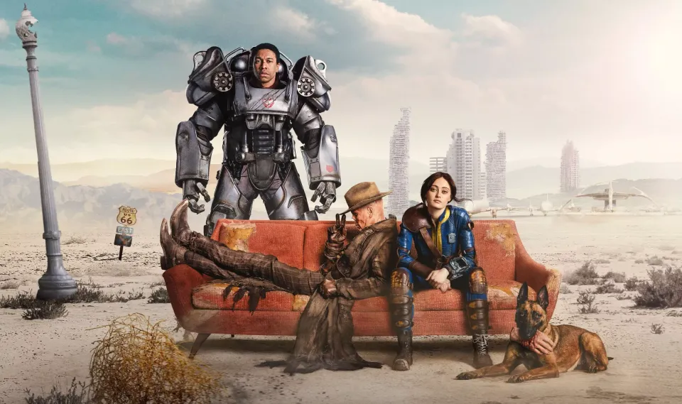 Lucy and the Ghoul sit on an orange couch, with Maximus in his power armor behind them. They're in the wasteland.