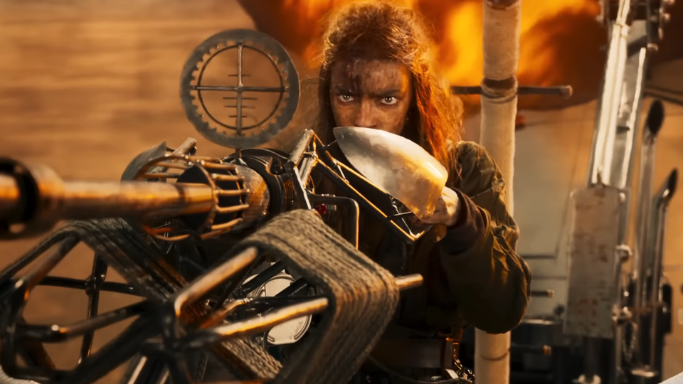 Furiosa, played by Anya Taylor-Joy, lines up a shot with an enormous gun. Fire belches behind her.