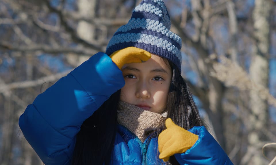 A young girl with long dark hair and brown eyes in a blue coat, grey hat, and yellow gloves walks in the forest.