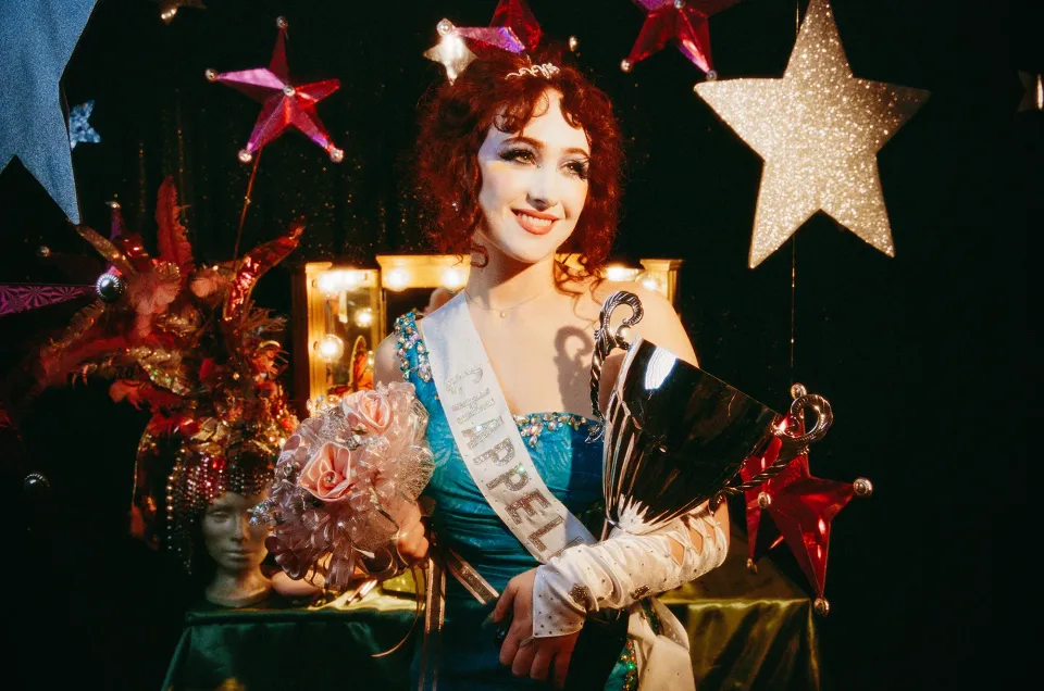 Pop star Chappell Roan wears a sash with her name on it, cradling flowers and a trophy.