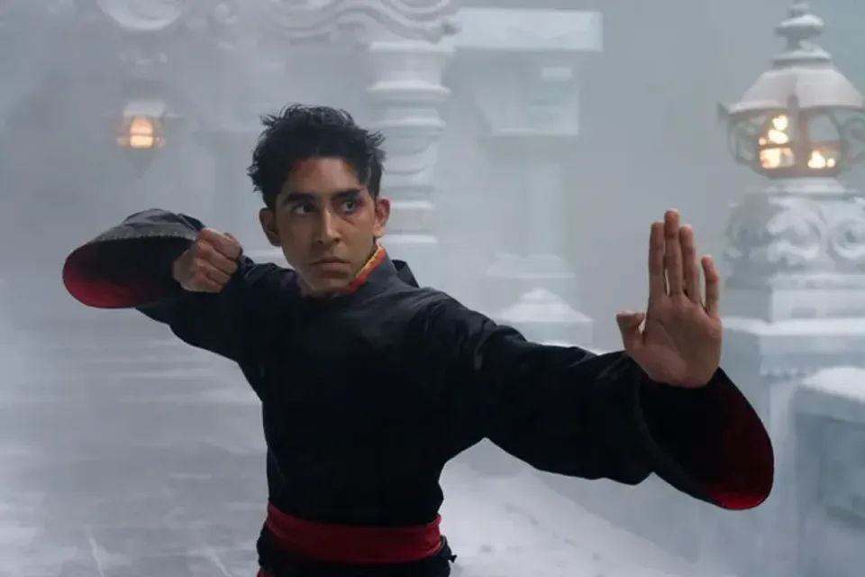 Dev Patel, dressed in a black and red robe, strikes a martial arts pose in a snowy temple.