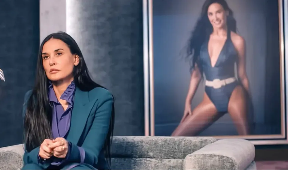 Our present-day Demi Moore sits on a couch right in front of a picture depicting her in the '80s.