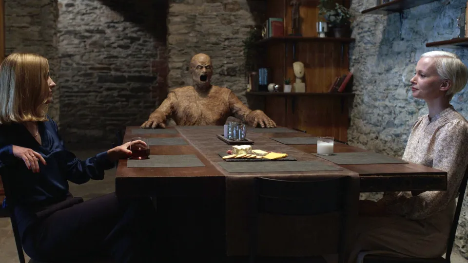 Two women sit at a long wooden table. At the other end of the table is a horrifying wooden man with mouth wide open.