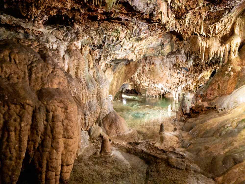 A cave is filled with brown stalactites and stalagmites. There's a pool of green water in the distance.